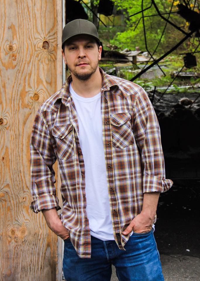 a man standing in front of a wooden wall with his hands in his pockets and looking at the camera