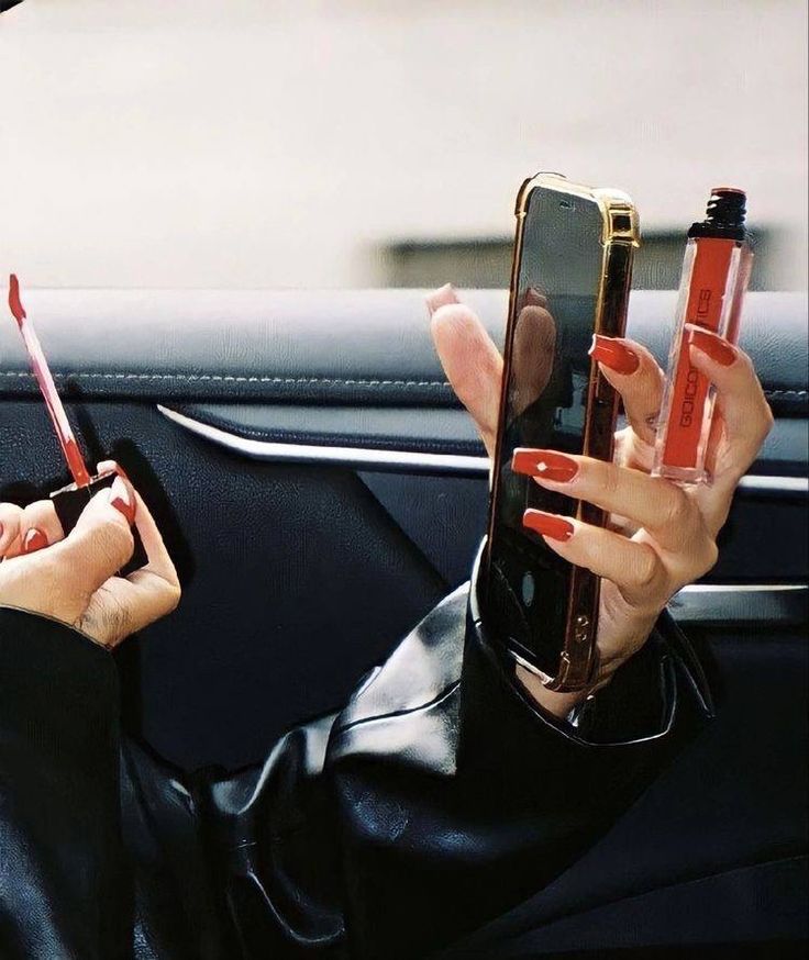 a woman sitting in the back seat of a car holding up her cell phone and lipstick