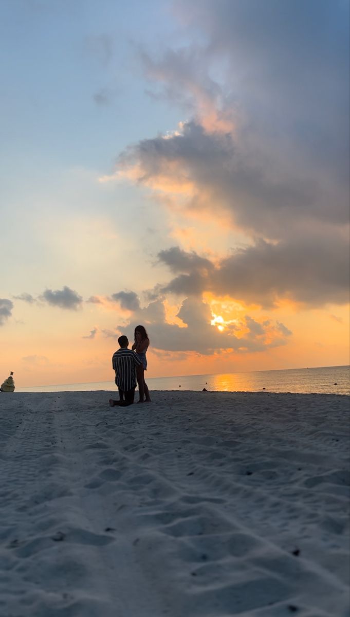 two people are sitting on the beach watching the sun rise over the water and clouds