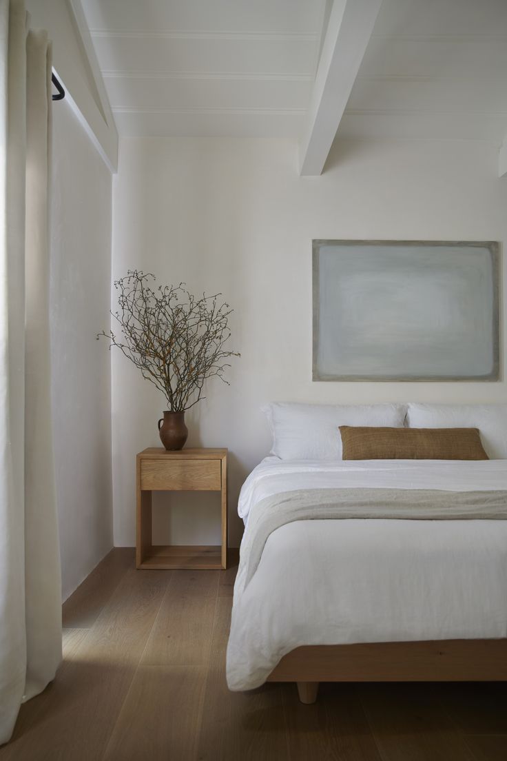 a white bed sitting next to a wooden table with a vase on top of it