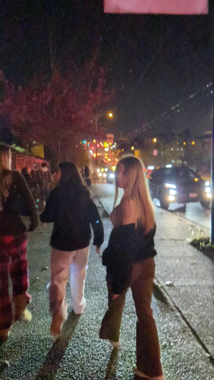 two women walking down the street at night with cars parked on the road behind them