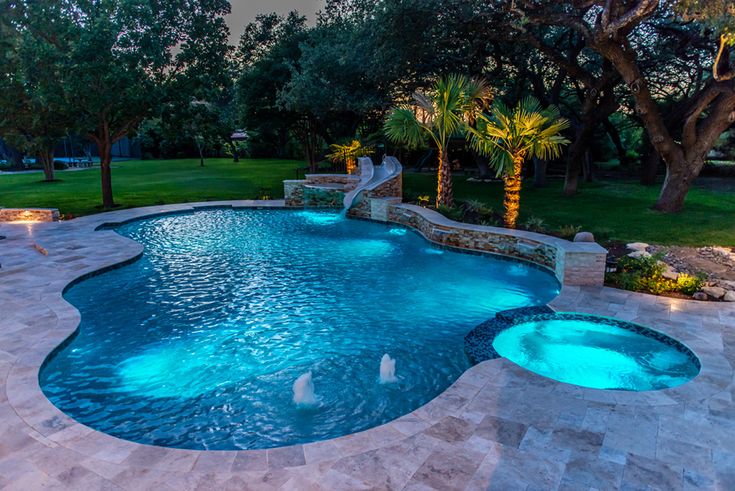 an outdoor swimming pool surrounded by landscaping and trees at night with lights on the water