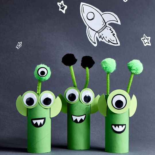 four green paper tubes with googly eyes and mouths in front of a chalkboard wall