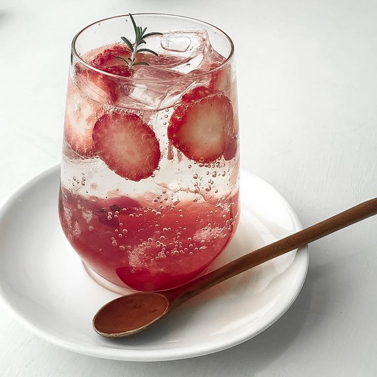 a glass filled with water and strawberries on top of a white plate next to a wooden spoon