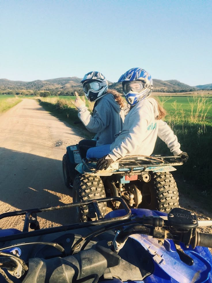 two people riding on the back of an atv down a dirt road next to a field