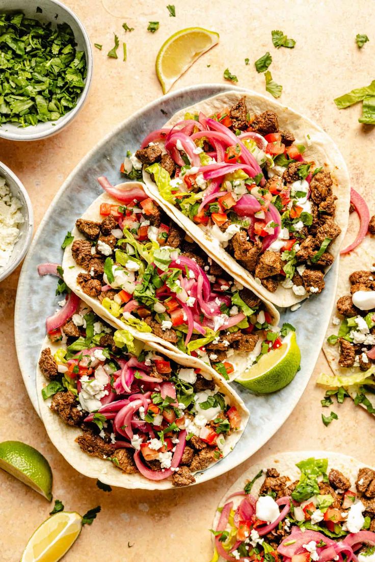 three tacos on a plate with limes, cilantro and other toppings