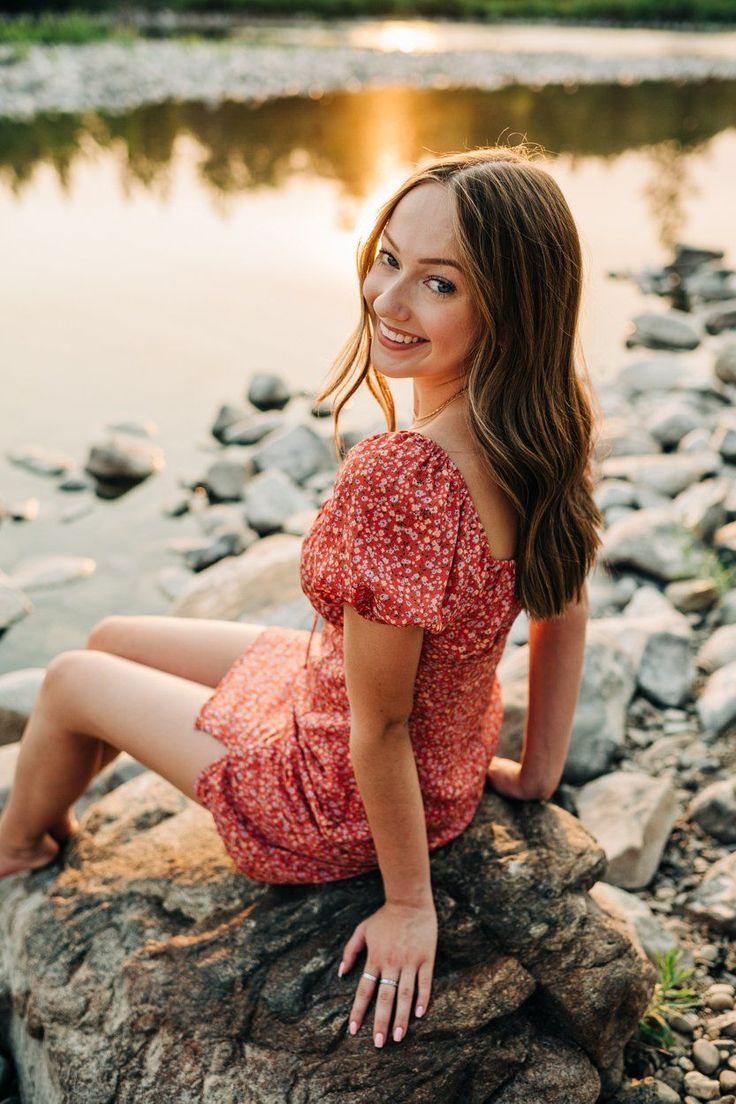 a beautiful young woman sitting on top of a rock next to a river at sunset