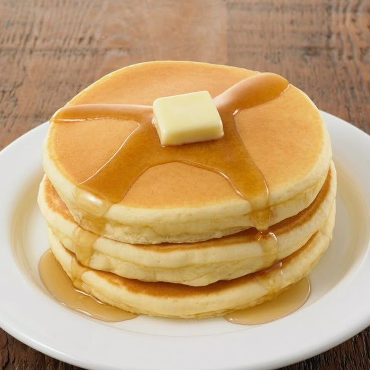 stack of pancakes with syrup and butter on top sitting on a white plate atop a wooden table