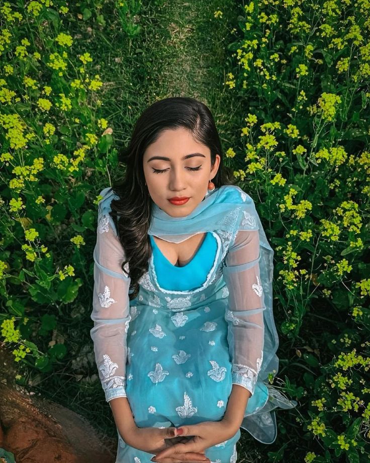 a woman standing in front of some flowers wearing a blue dress and holding her hands together