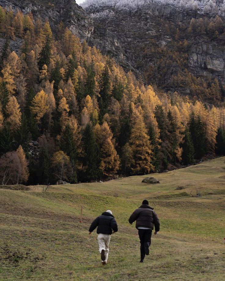 two people are running through the grass in front of some trees with yellow leaves on them