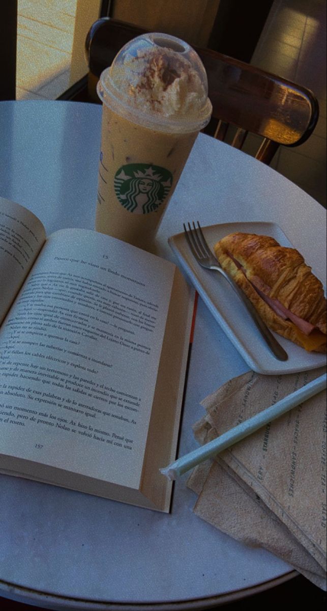 an open book sitting on top of a table next to a cup of coffee and croissant