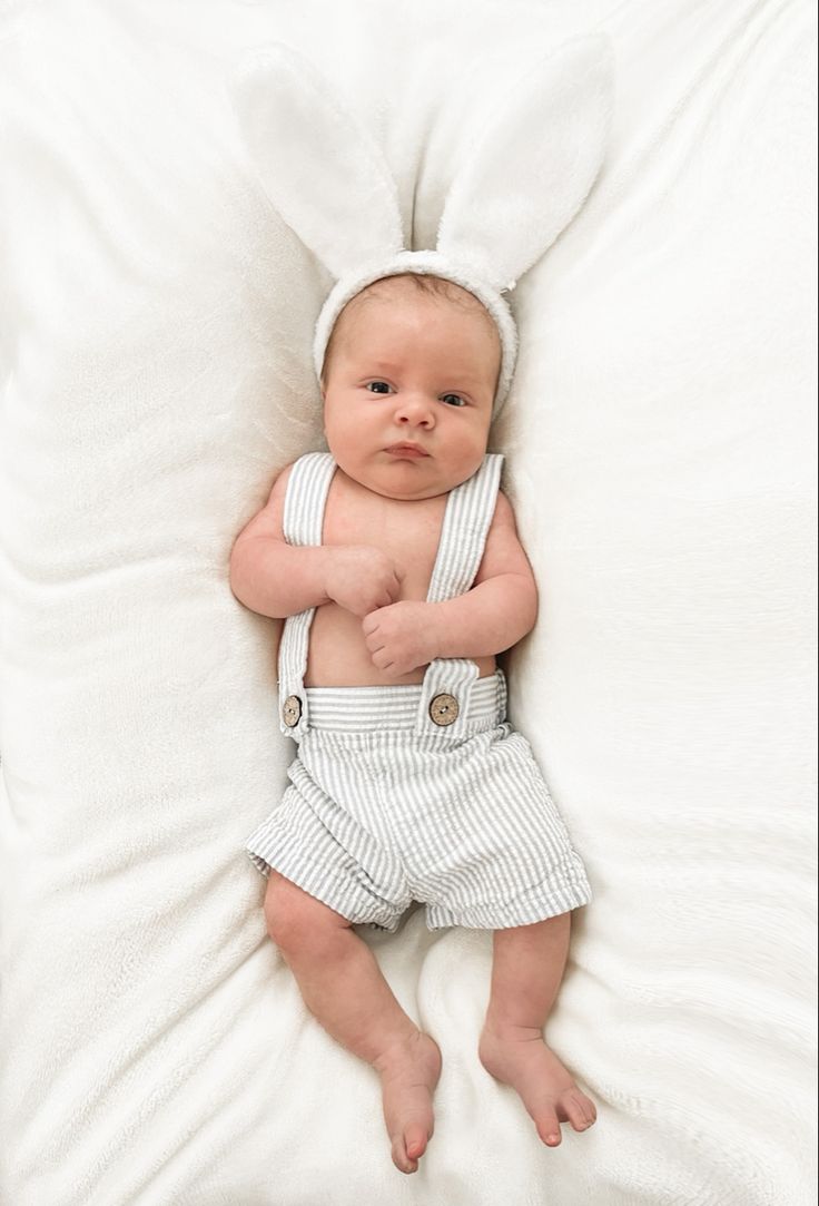 a baby in overalls laying on top of a white blanket with his arms wrapped around him