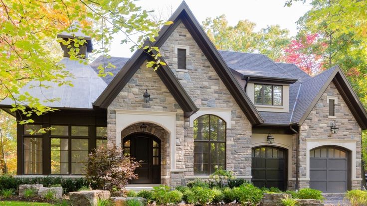 a stone house with two garages and landscaping in the front yard, surrounded by trees