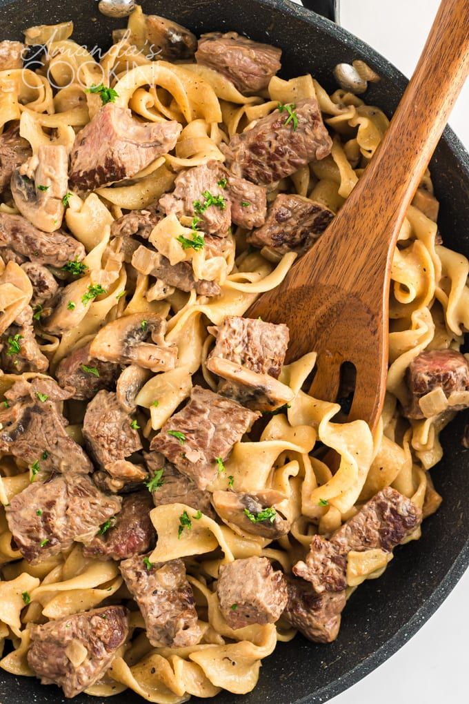 a skillet filled with pasta and meat on top of a white table next to a wooden spoon