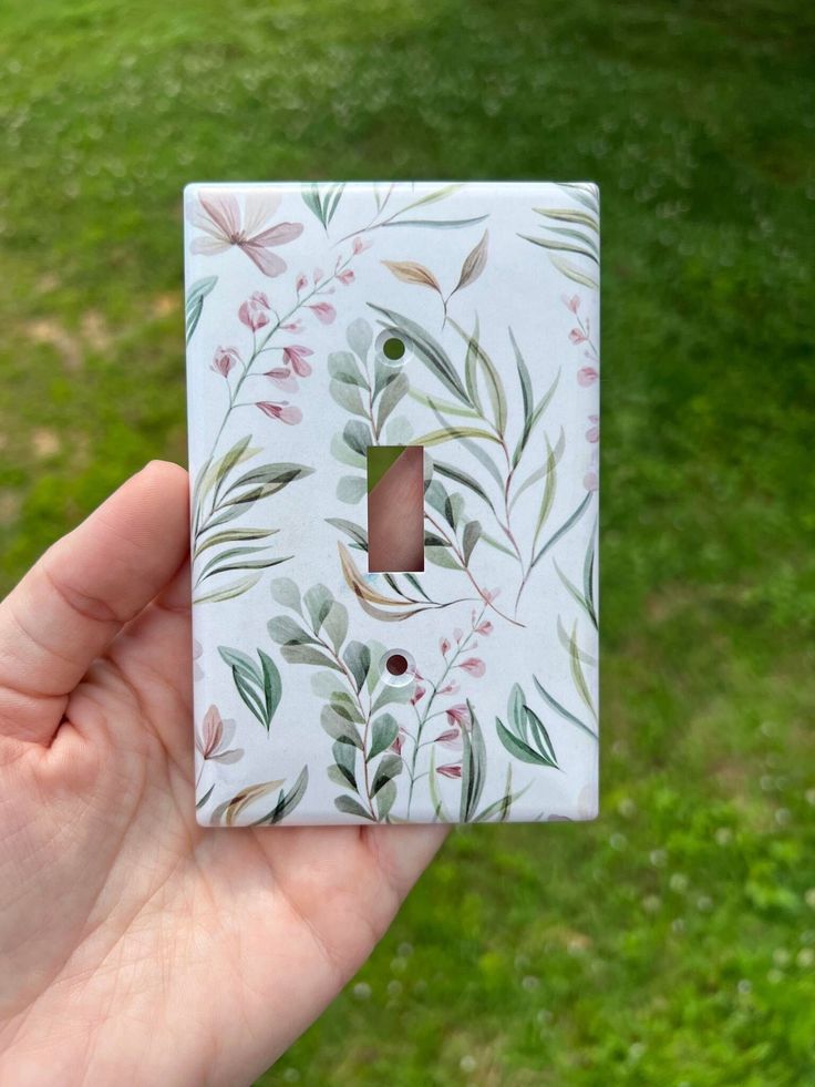 a hand holding a light switch cover in front of a green field with grass and flowers