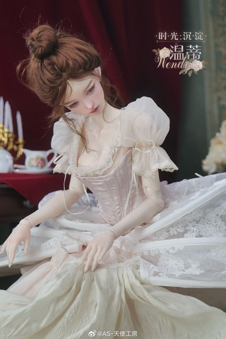 the doll is wearing a white dress and holding her hands out to her chest, while sitting on a table