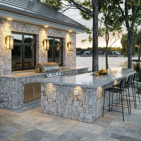 an outdoor kitchen with stone counter tops and bar stools next to the water at sunset