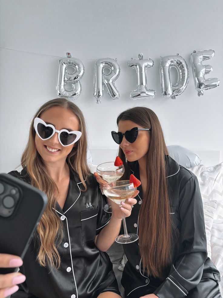 two women sitting on a bed holding glasses and looking at a cell phone in front of them