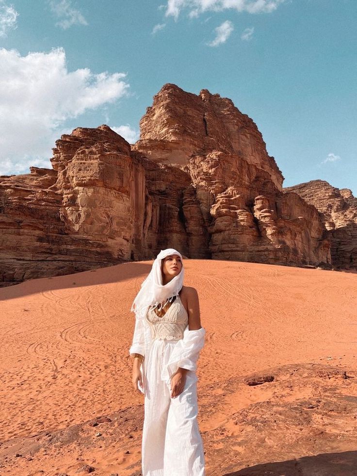 a woman standing in the desert wearing a white dress and headdress with mountains in the background