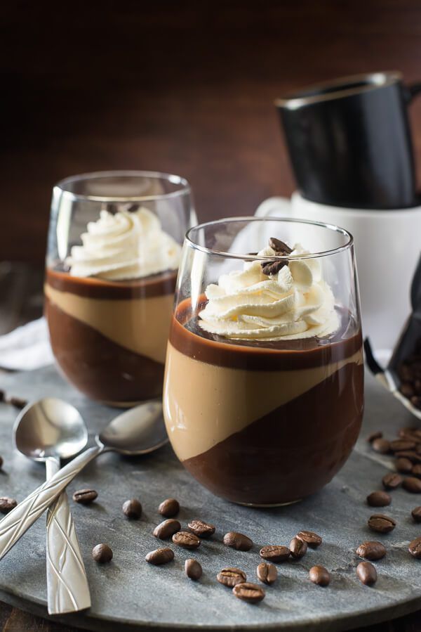 two glasses filled with dessert sitting on top of a table next to coffee beans and spoons