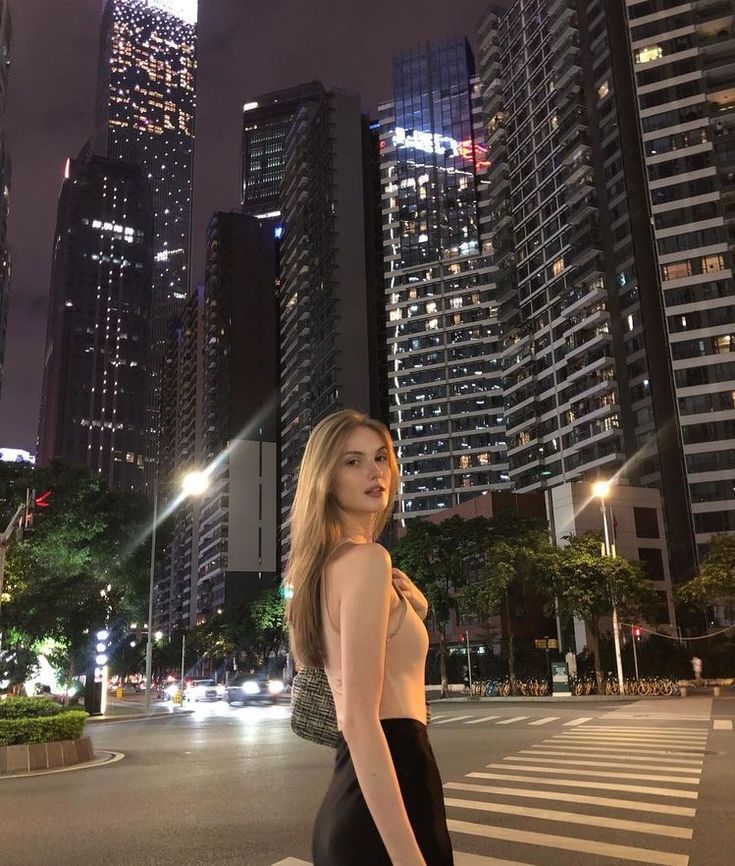 a woman standing in the middle of a street at night with tall buildings behind her