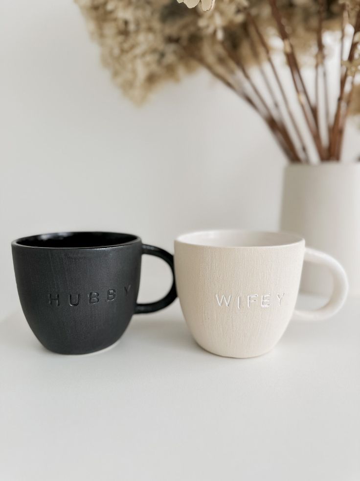 two black and white mugs sitting next to each other on a table with dried flowers in the background