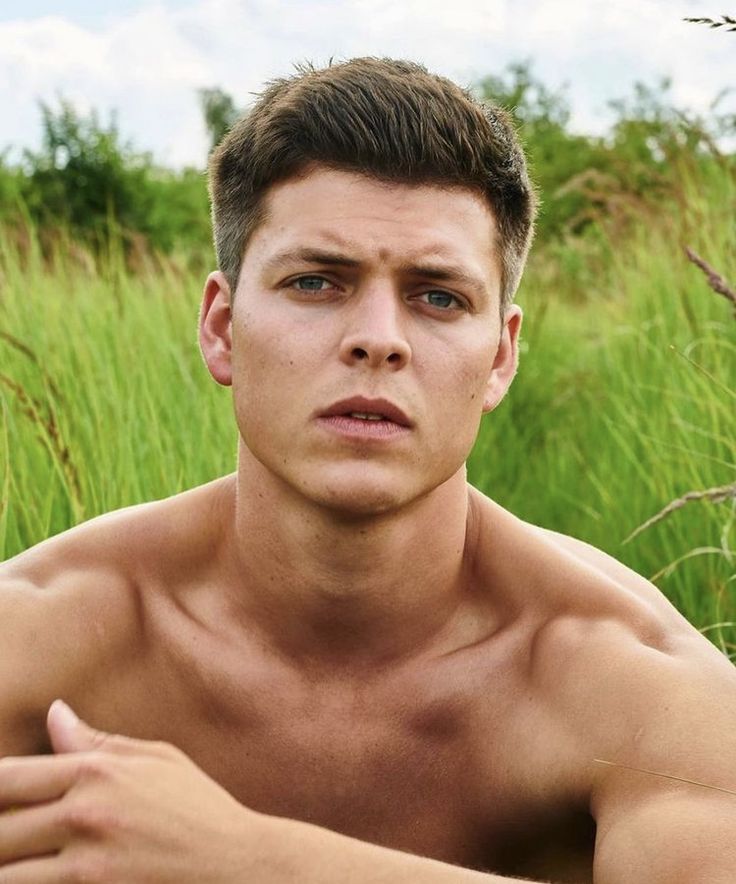 a shirtless man sitting in tall grass with his hand on his chest looking at the camera