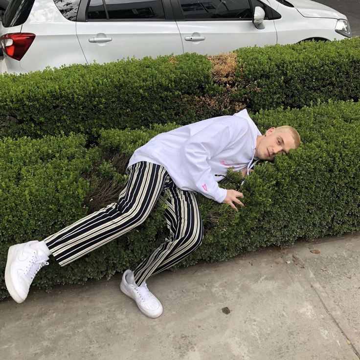 a man in striped pants leaning against a hedge next to a white car on the street