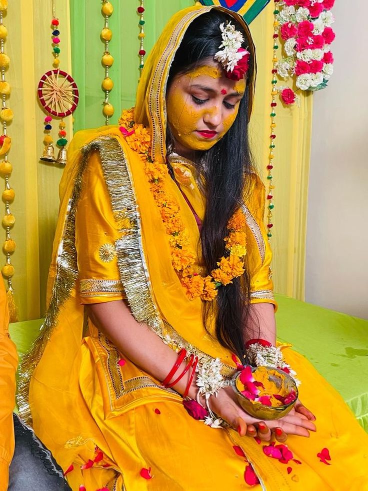 a woman dressed in yellow sitting on a bed with flowers around her head and holding a bowl