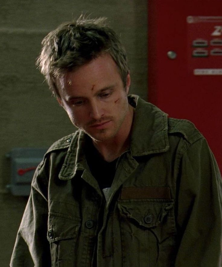 a man in an army green jacket looking down at his cell phone while standing next to a red door