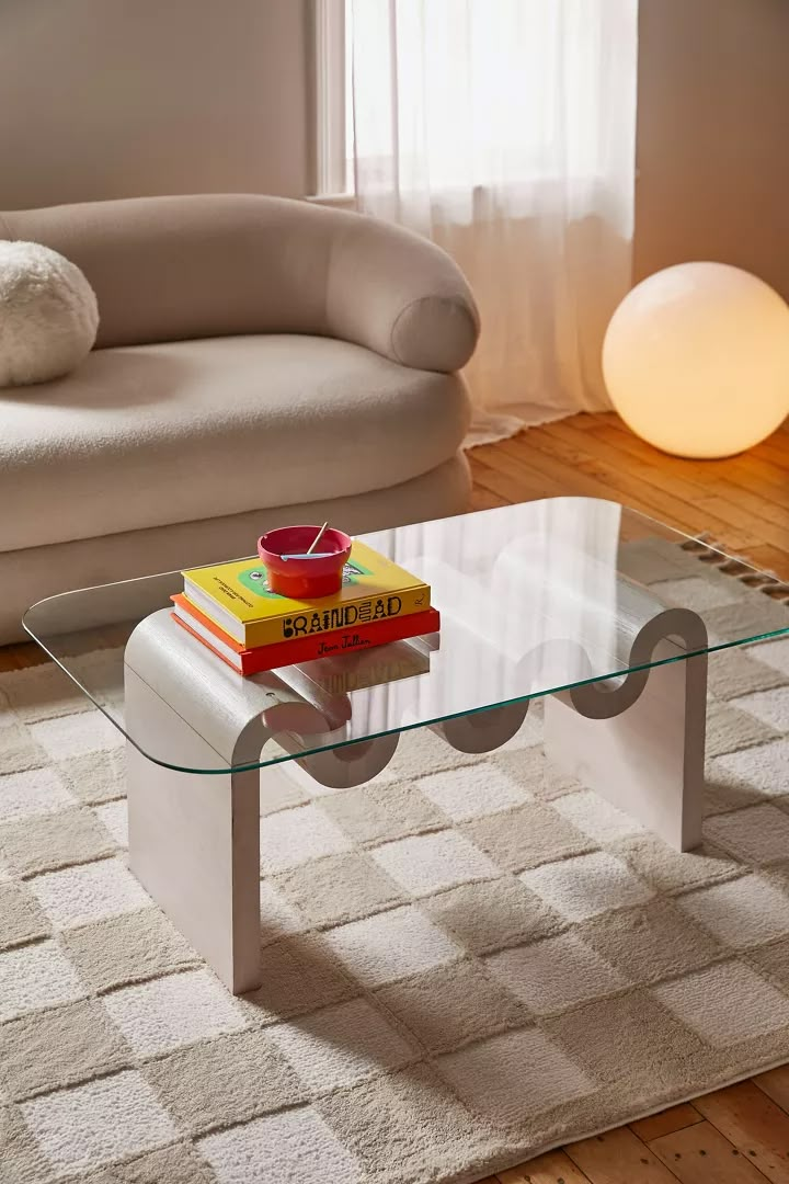 a glass table with books on it in front of a white couch and chair next to a window