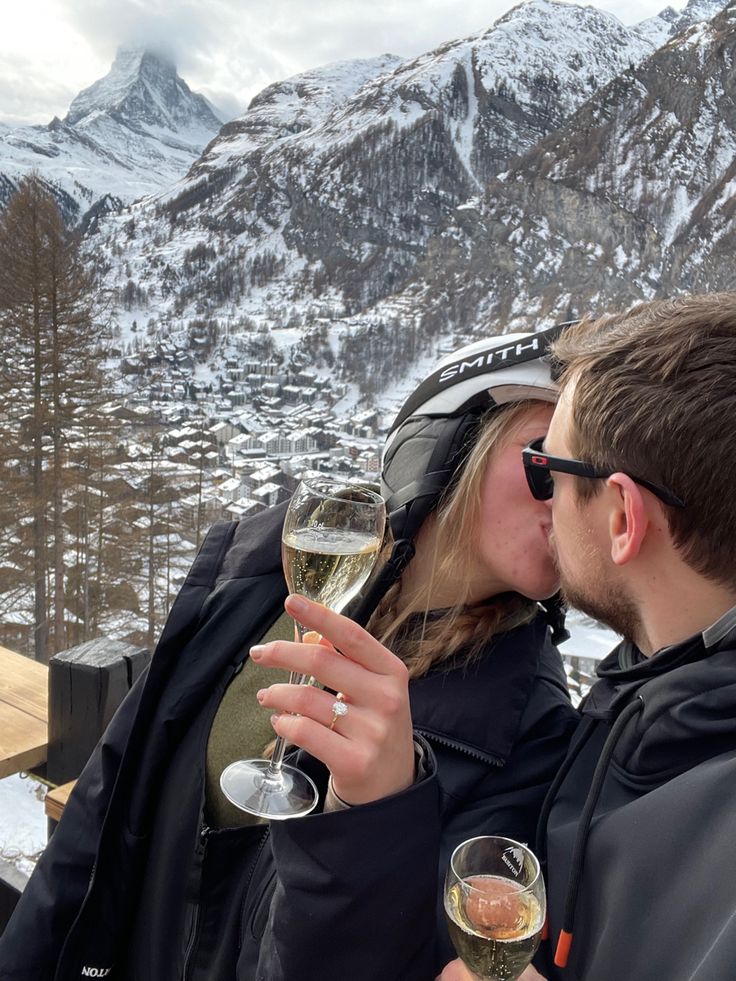 a man and woman kissing while holding wine glasses in front of snow covered mountain range