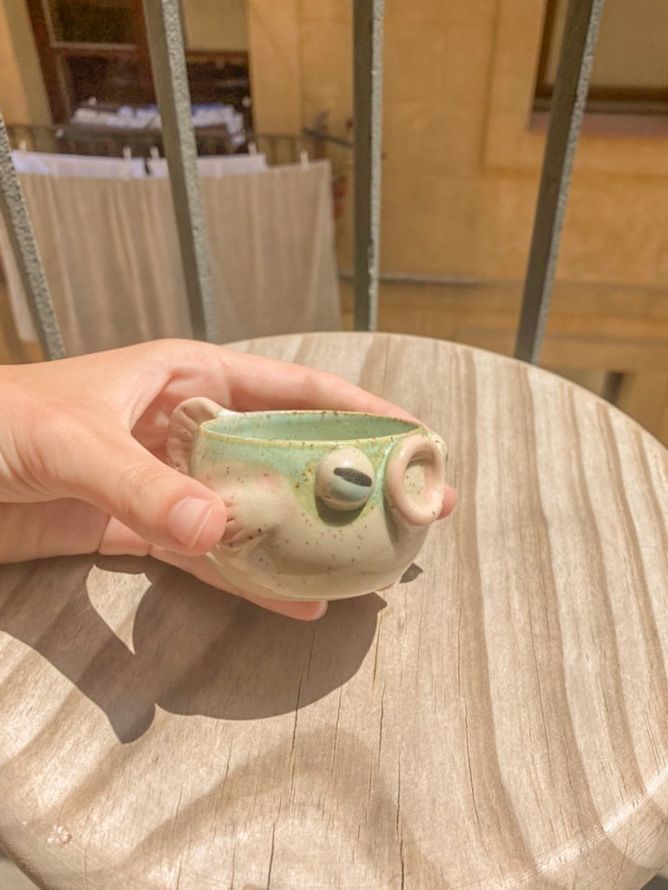 a hand holding a small ceramic cup on top of a wooden table with chairs in the background