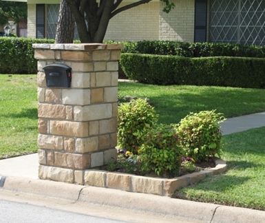 a brick mailbox in front of a tree and bushes on the side of a street