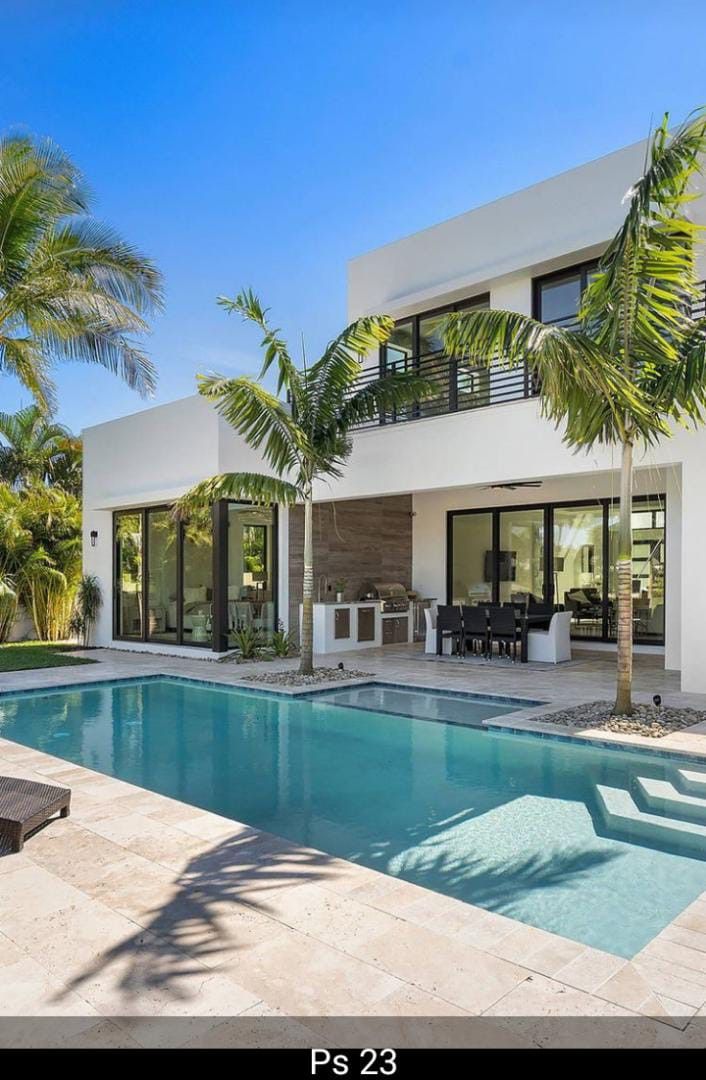 a large swimming pool in front of a white house with palm trees and chairs around it