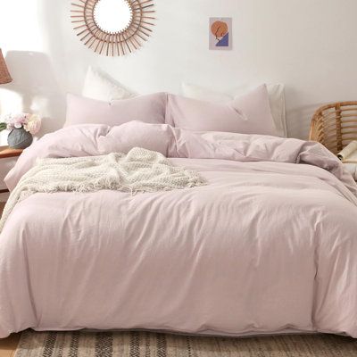 a bed with pink comforter and pillows in a white room next to a wicker chair