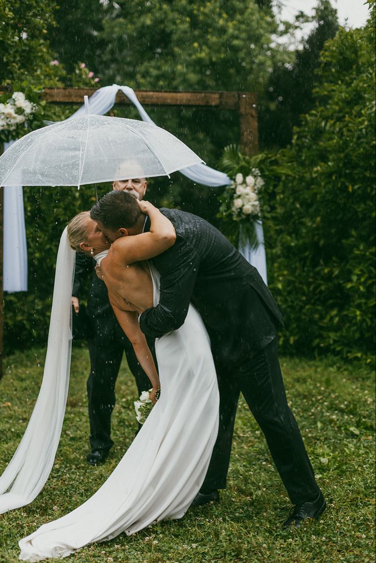 a bride and groom hug under an umbrella in the rain at their outdoor wedding ceremony