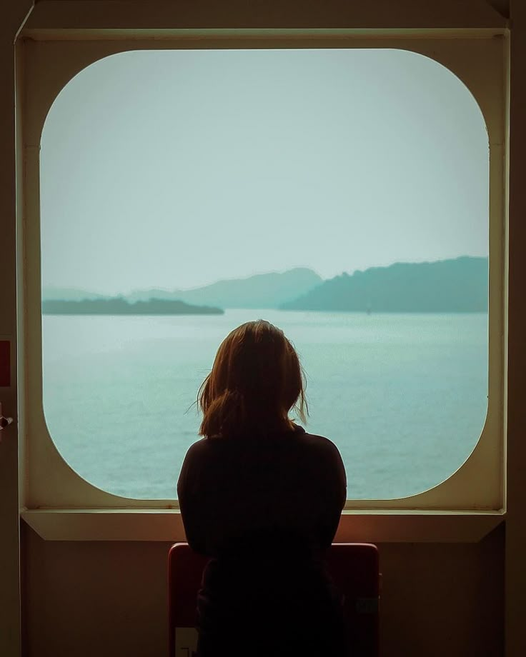 a woman sitting in front of a window looking out at the water