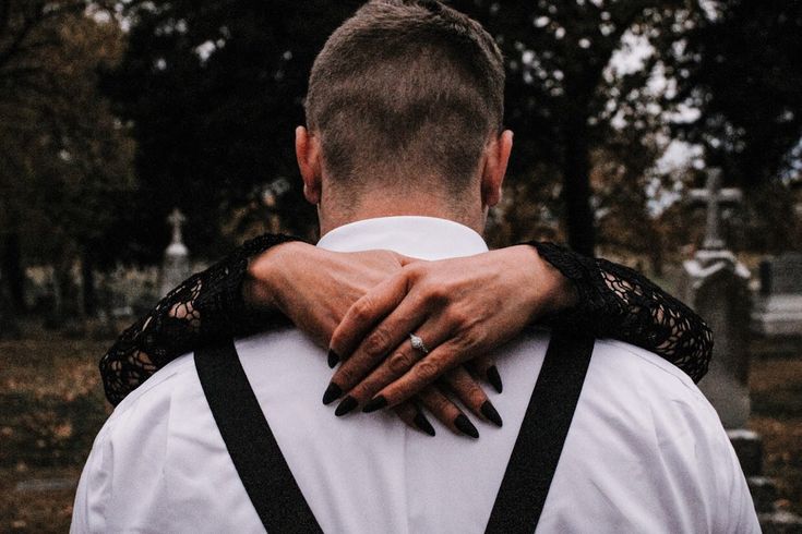 a man in white shirt and black suspenders hugging another man's shoulder at cemetery