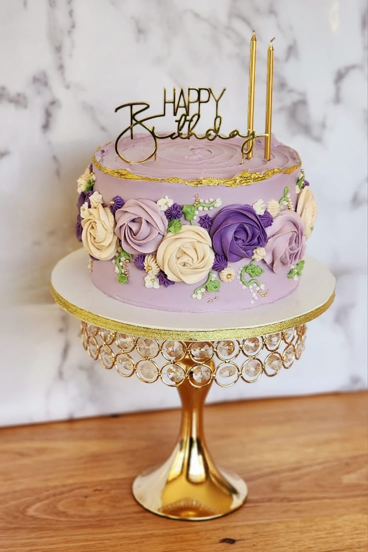 a birthday cake decorated with purple flowers and gold trimmings on a stand in front of a marble wall