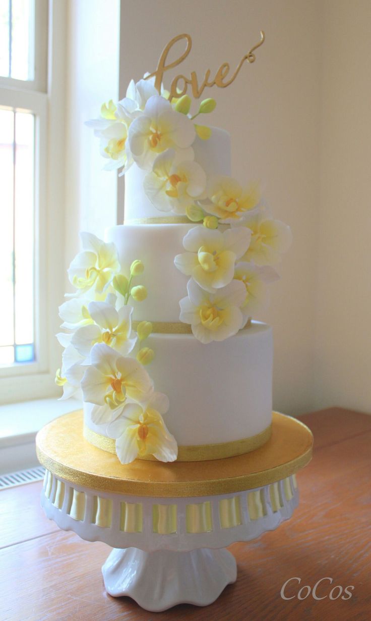 a three tiered cake with white and yellow flowers on the top, in front of a window