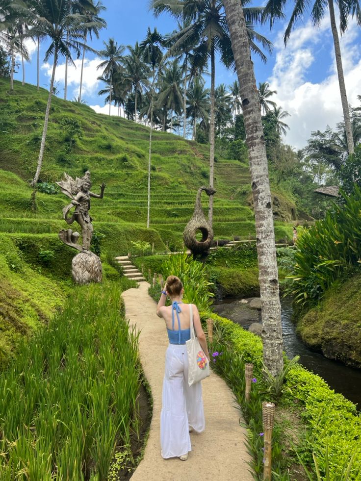 a woman walking down a path next to lush green hillside covered in trees and palm trees