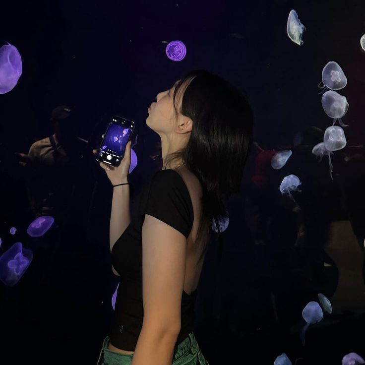 a woman holding a cell phone in front of jelly fish floating around her and looking up at the sky