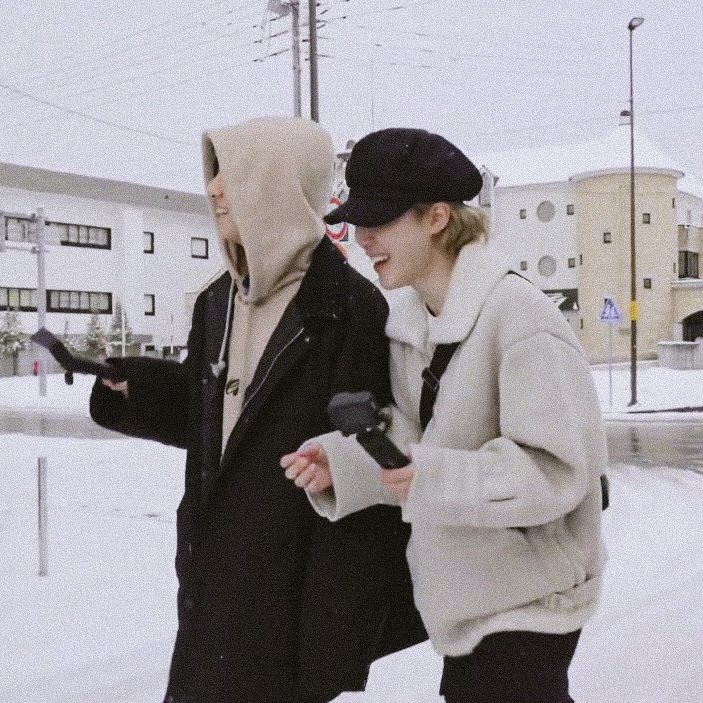 two people walking in the snow with one wearing a hood and another holding a cell phone