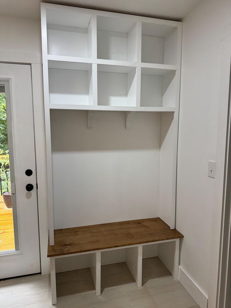 an empty room with white shelving and wooden bench in the corner, next to a glass door