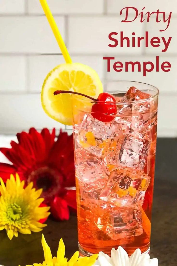 a drink in a tall glass with ice and a cherry on the rim next to flowers