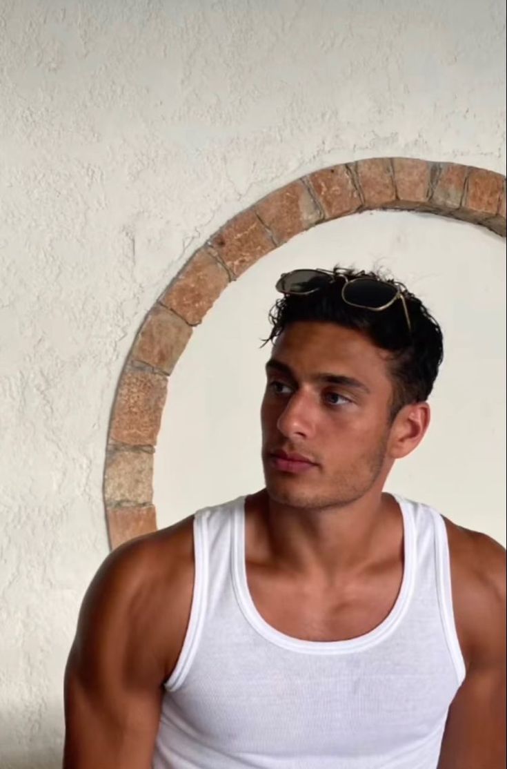 a man in white tank top standing next to a brick wall and looking at the camera