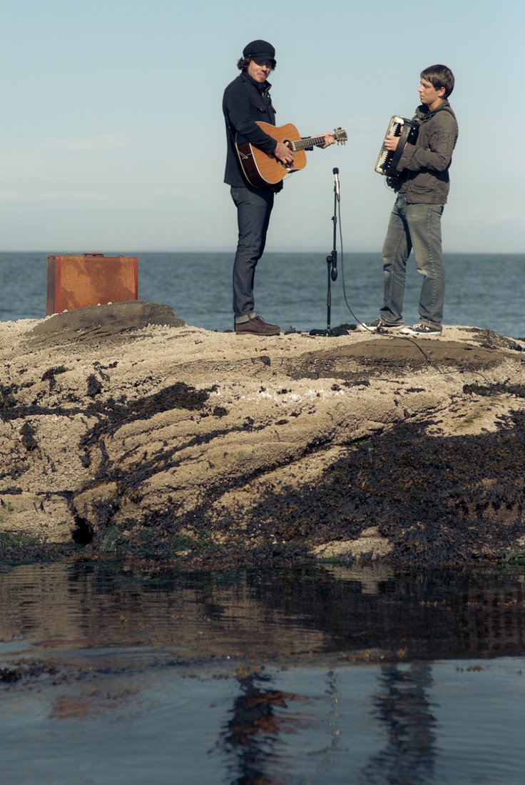 two men standing on top of a rock next to the ocean playing guitar and singing