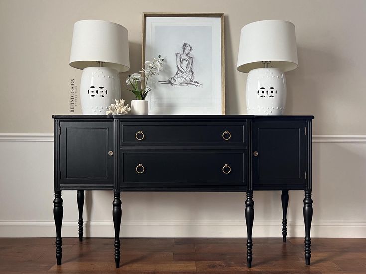 two lamps are sitting on top of a black dresser next to a white vase with flowers