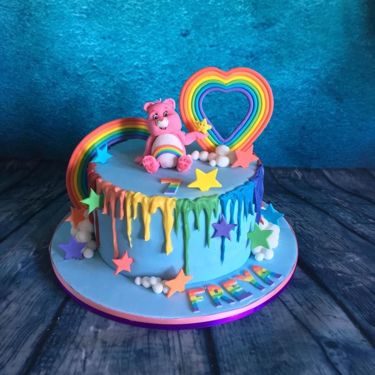 a blue cake with rainbow icing and a pink teddy bear on top, sitting on a wooden table